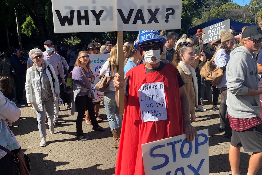 Un homme masqué et portant une cape rouge tient une pancarte disant « arrêtez le chantage vax ».